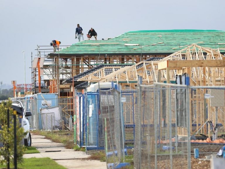 Construction site with multiple workers on scaffolding and the roof of a partially built house, surrounded by fencing and construction materials